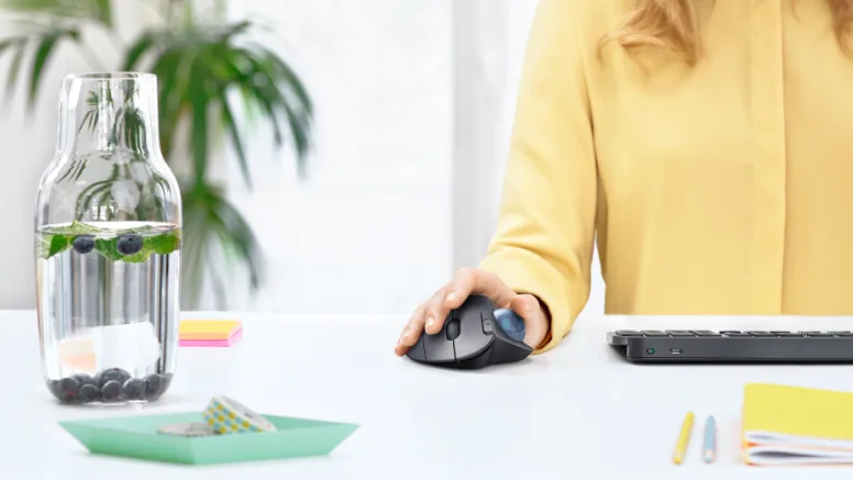 Person in yellow shirt working at desk using ergonomic mouse and keyboard in bright office environment