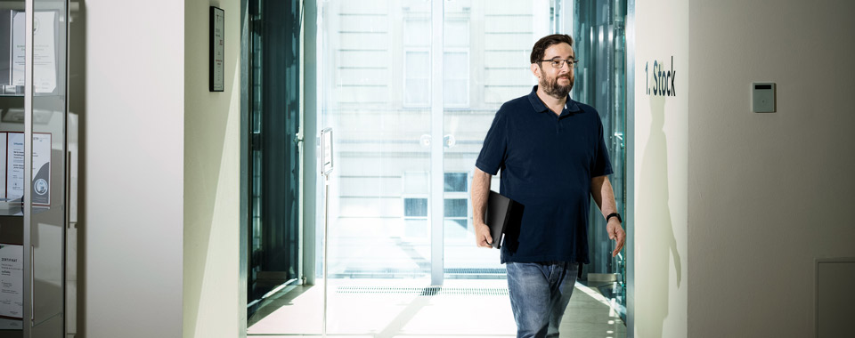 Mann mit Laptop in der Hand geht durch hellen Flur mit großen Fenstern in moderner Arbeitsumgebung