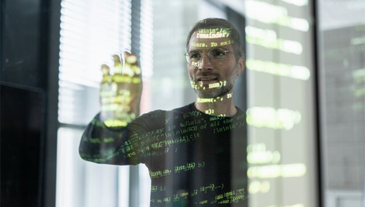 Man wearing glasses focused on programming code displayed on a transparent screen in a modern office