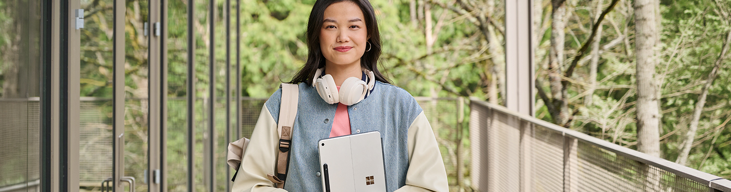 A young woman with a Microsoft Surface tablet, headphones around her neck, and a backpack, standing on a grassy sidewalk outdoors.