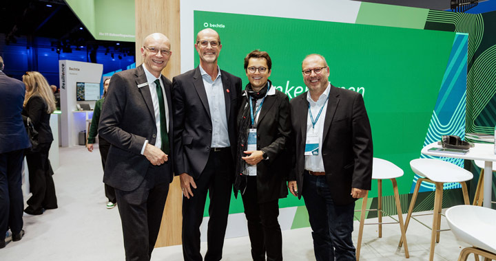 Vier Personen in Businesskleidung auf einer Messe vor grünem Messestand mit Firmenbranding
