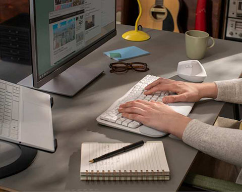 Person typing on keyboard at desk with monitor, notebook, and coffee cup in work environment
