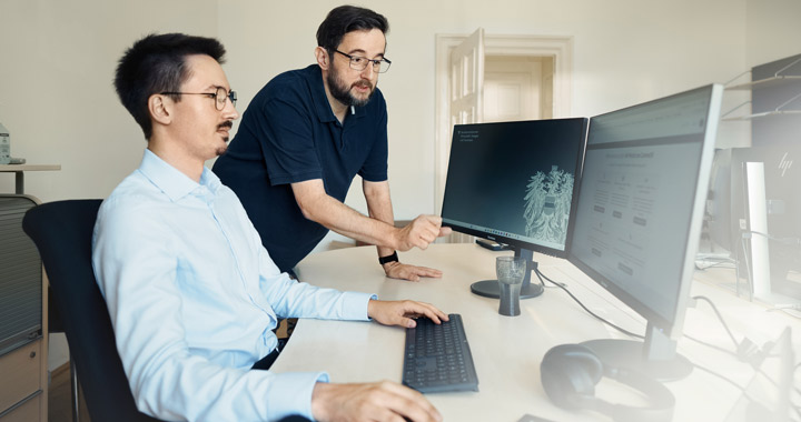 Zwei Personen im Büro bei gemeinsamer Arbeit an Computern mit Fokus auf Bildschirm und Zusammenarbeit