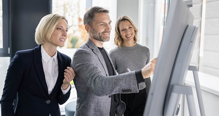 Zwei Frauen und ein Mann treffen sich vor einer digitalen Anzeigetafel.