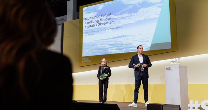 Zwei Personen präsentieren auf einer Bühne vor Publikum ein digitales Ökosystem für Multicloud-Lösungen