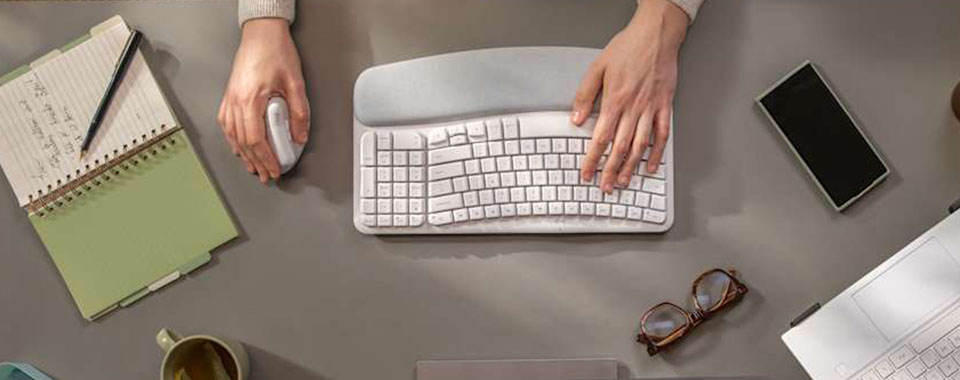 Person working at desk with ergonomic keyboard and mouse surrounded by notebook, smartphone, and glasses