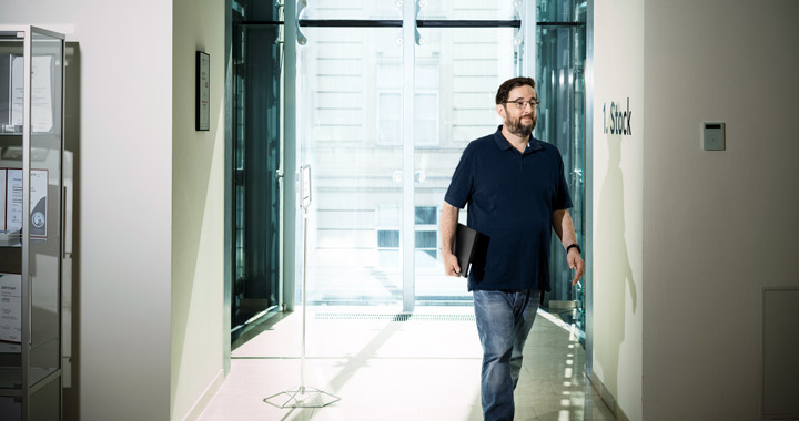 Mann mit Laptop in der Hand geht durch hellen Flur mit großen Fenstern in moderner Arbeitsumgebung
