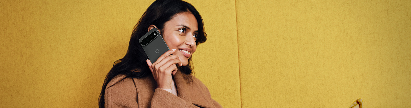 Woman in brown coat smiling while talking on google pixel 11 smartphone against yellow background
