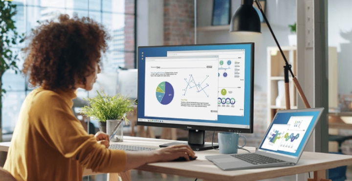 Woman working at desk with monitor and laptop displaying charts