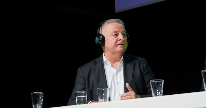 Mann mit Headset spricht bei Podiumsdiskussion vor dunklem Hintergrund und mehreren Wassergläsern