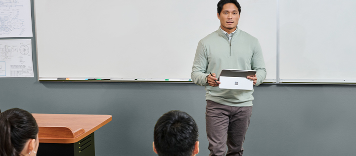 A teacher stands in front of a class with a Microsoft Surface device, teaching students in a modern classroom.