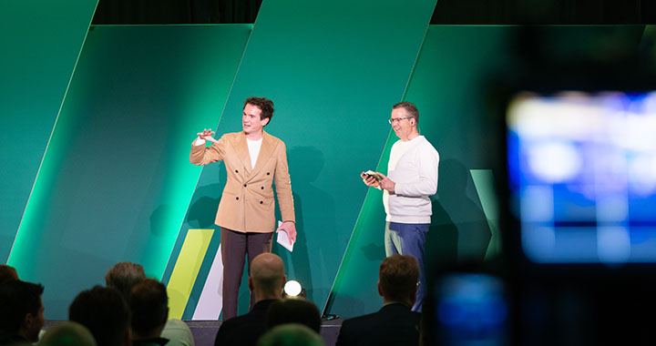 Two people on stage presenting to an audience in a modern event space with green backdrop