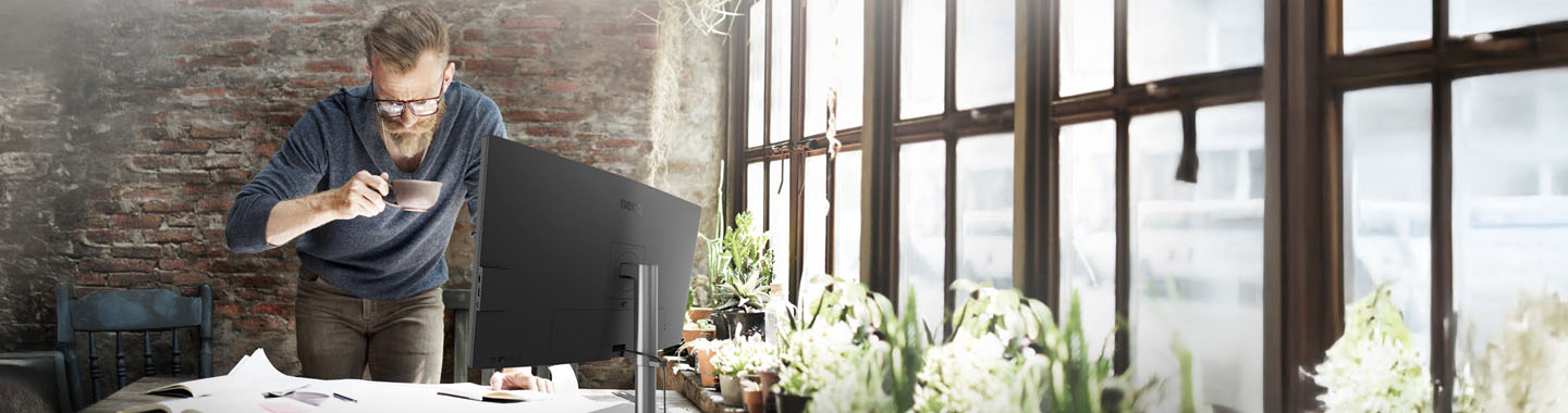 Man holding a coffee cup in an office with large windows. There is a large monitor on the desk and many plans are spread out on it.