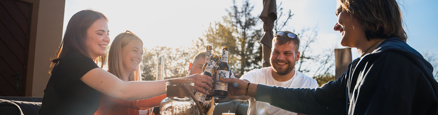 A group of five people are sitting outside at a table in the lovely weather, clinking their beer bottles together.